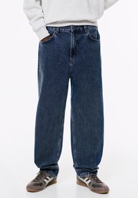 Jeans larghi blu scuro realizzati in denim, con un classico design a cinque tasche e una finitura leggermente sbiadita. Abbinati a scarpe da ginnastica grigie.