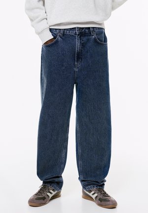 Jeans larghi blu scuro realizzati in denim, con un classico design a cinque tasche e una finitura leggermente sbiadita. Abbinati a scarpe da ginnastica grigie.