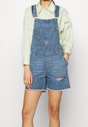 Kvinde iført blå denim kort overall med en forlomme og en lysegrøn skjorte med lange ærmer og knapper.