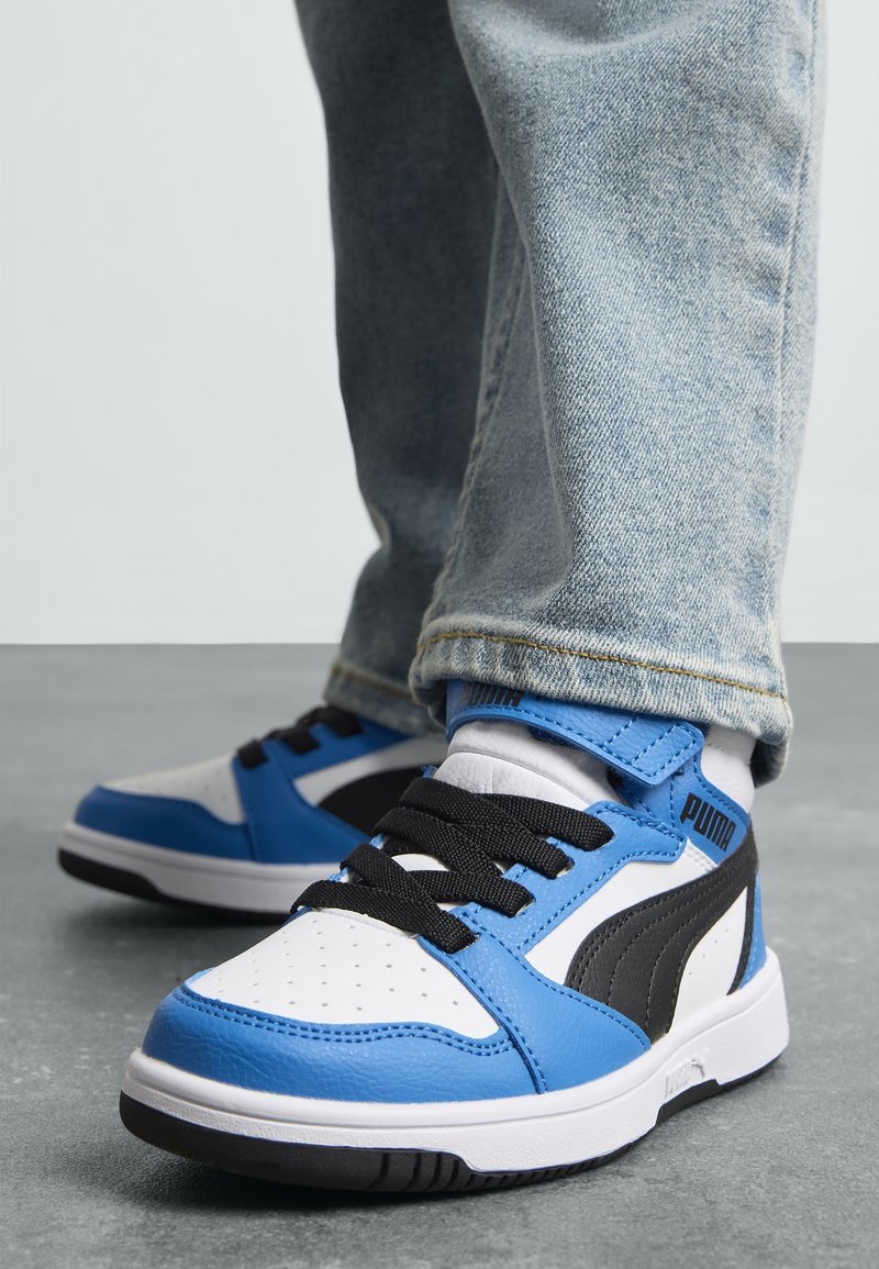 Personne portant des baskets Puma bleu clair et blanc avec des lacets noirs et un jean, debout sur un sol gris.