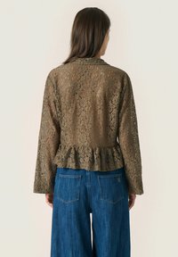 Lace blouse with long sleeves, olive green, featuring a peplum hem and intricate floral patterns. Paired with wide-leg denim jeans.