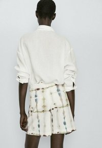 Chemise grande taille en lin blanc à manches retroussées, associée à un short crème présentant un motif tie-dye aux tons verts, marron et bleus.