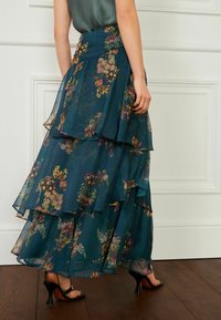 Teal lagd nederdel med blomstermønster, der har gennemsigtig stof og flæser i kanten. Bæres med sorte åben-tå hæle. Gulvlængde design.