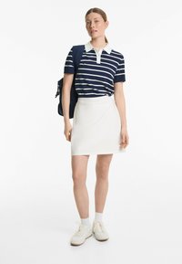 Polo rayé bleu marine et blanc à manches courtes, associé à une jupe blanche. Le modèle porte des baskets blanches et tient un sac bleu.