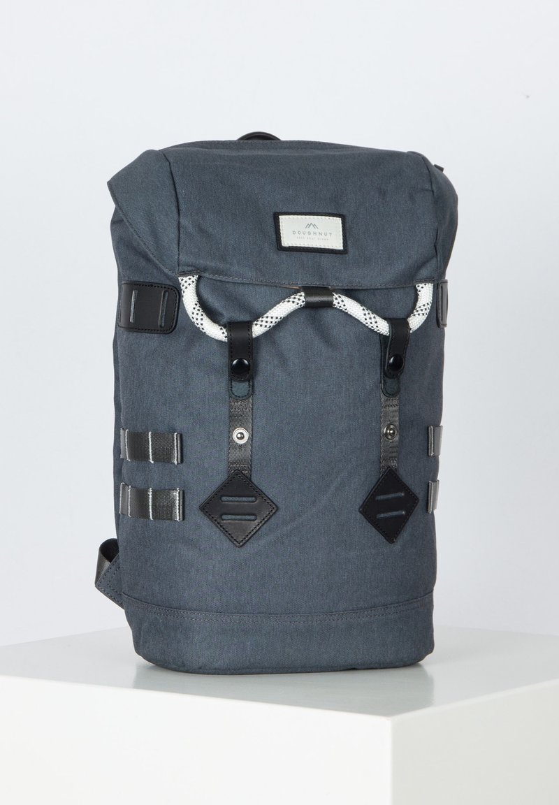 Sac à dos en tissu gris avec un design à rabat, accents noirs externes et fermeture à cordon à motif. Comprend des sangles latérales et un patch logo.