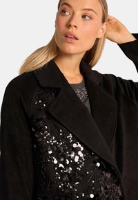 Cappotto nero con un design di paillettes sul lato sinistro, texture liscia e colletto a risvolto. Sotto c'è una maglia grigia con decorazioni.