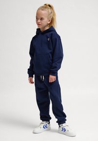 Marinblå hoodie med ficka fram och liten emblem, tillsammans med matchande joggerbyxor och vita sneakers med blå accenter.