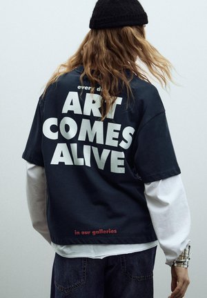Person med langt hår iført en sort beanie og lagdelte sorte og hvide skjorter med teksten "KUNSTEN BLIVER LEVENDE i vores gallerier" trykt på bagsiden.