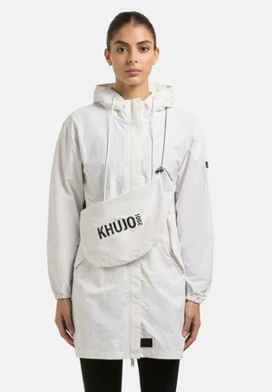 Vrouw draagt een wit jack met capuchon en een crossbodytas met het logo "KHUJO 2001" aan de voorkant, staand tegen een effen witte achtergrond.