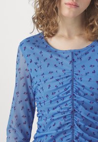 Femme portant un haut bleu transparent à manches longues avec un petit imprimé floral violet et des détails froncés sur le devant au centre.