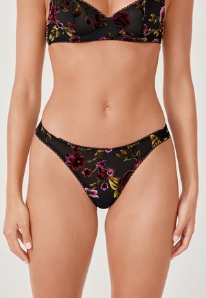 Schwarzes, durchsichtiges Lingerie-Set mit bestickten, mehrfarbigen Blumenmustern, getragen von einer stehenden Frau vor einem schlichten Hintergrund.