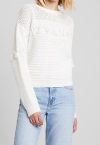 Weißer Strickpullover mit offenem Strickmuster, strukturierten Streifen und ausgefransten Akzenten an den Ärmeln. Kombiniert mit hellblauen Jeans.