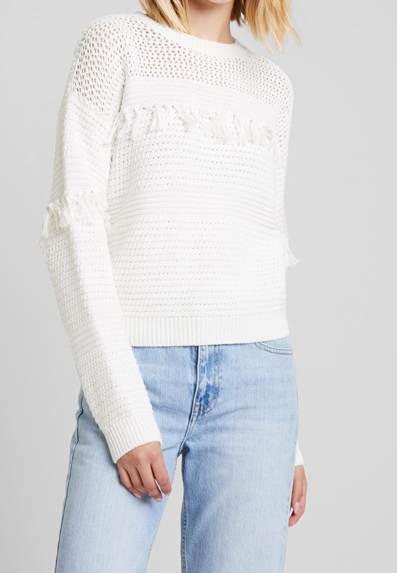 Weißer Strickpullover mit offenem Strickmuster, strukturierten Streifen und ausgefransten Akzenten an den Ärmeln. Kombiniert mit hellblauen Jeans.