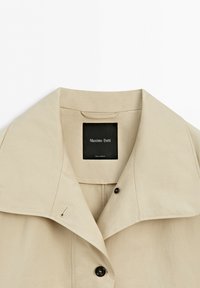 Beżowy kołnierz płaszcza trenczowego z szerokimi klapami i czarnymi guzikami, z widoczną metką marki „Massimo Dutti” oraz metką „Made in Morocco” wewnątrz.