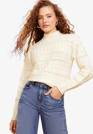 Kermanvärinen neulepaita, jossa on ribbauskaulus ja teksturoitu pinta. Yhdistetty korkeavyötäröisiin sinisiin farkkuihin, joissa on klassinen denim-ilme.