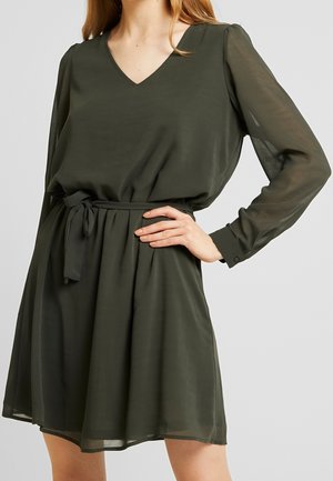 Femme portant une robe courte en voile vert foncé à manches longues avec un col en V et une ceinture nouée à la taille, main posée sur la hanche.
