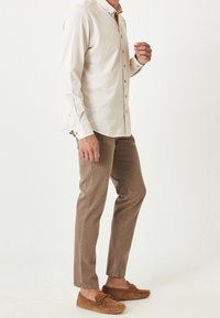 Camisa de botones de manga larga color beige con cuello, combinada con pantalones ajustados de color marrón claro y zapatos mocasín de ante marrón. Diseño limpio y minimalista.