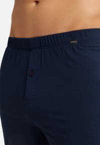 Pantaloni lounge blu navy in tessuto morbido. Presentano una vita elasticizzata, chiusura con bottone frontale e un piccolo etichetta del marchio. Design classico e funzionale.