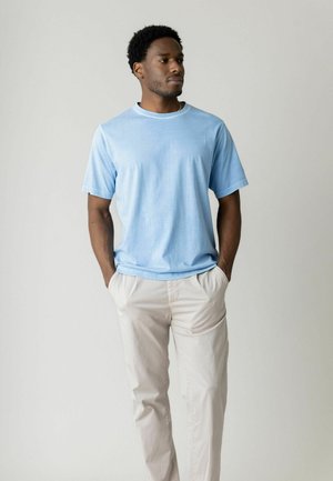 Muž v svetlomodrom tričku a béžových nohaviciach stojí s rukami v vreckách, pozerá sa do strany na jednoduchom svetlosivom pozadí.