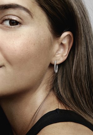 Silberner Creolenohrring mit strukturiertem, gedrehtem Design. Das Modell hat lange braune Haare und trägt ein schwarzes Oberteil. Nahaufnahme des Ohrs und des Ohrrings.
