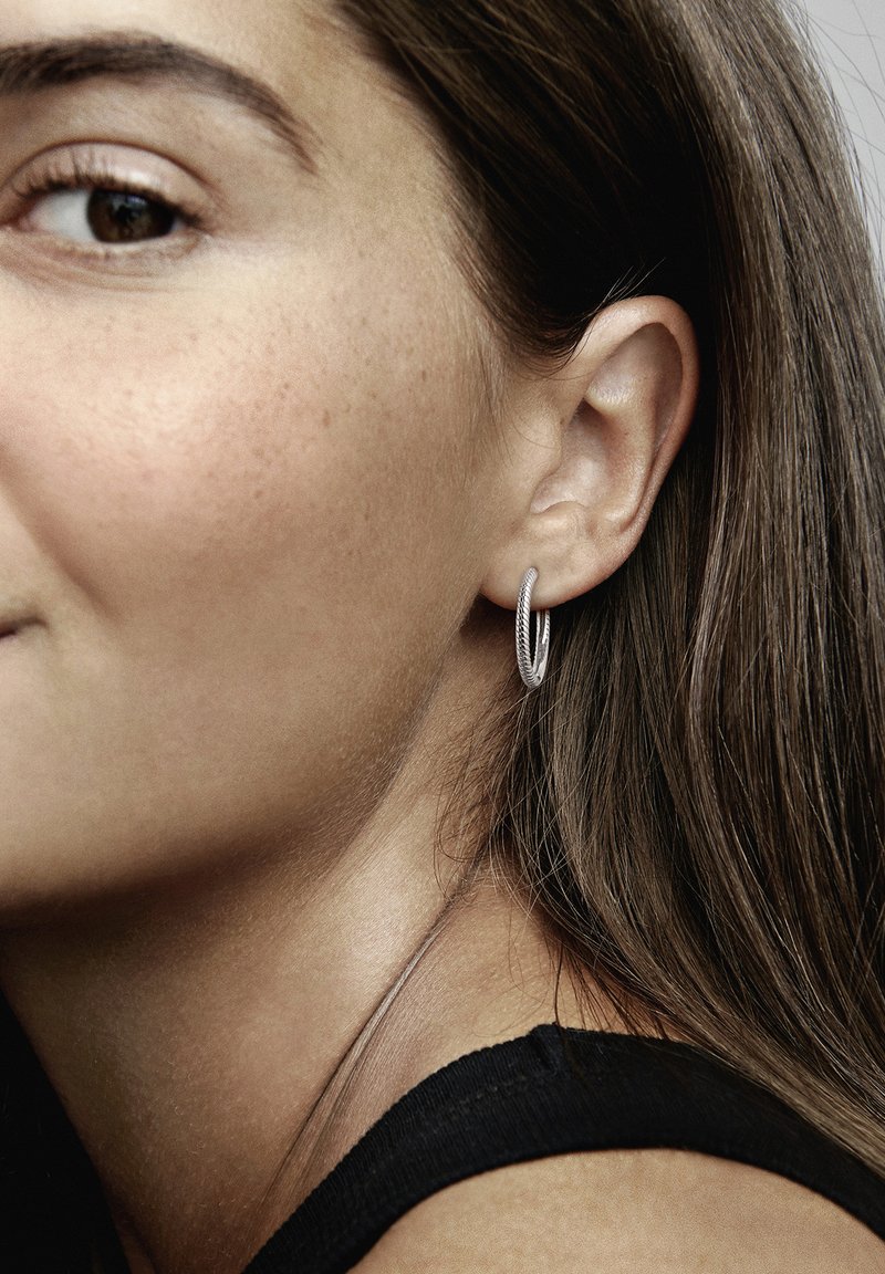 Pendiente de aro de plata con un diseño texturizado y retorcido. El modelo tiene el cabello largo y castaño, y lleva una blusa negra. Vista en primer plano de la oreja y el pendiente.