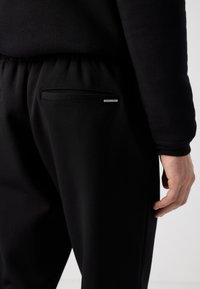 Black pants featuring a smooth texture, elastic waistband, and two back pockets with subtle hardware detailing.