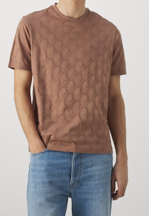 Mann trägt braunes, gemustertes Kurzarm-T-Shirt und blaue Jeans, eine Hand in der Hosentasche vor einem schlichten hellen Hintergrund.