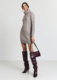 Vestito color beige chiaro a maglia con collo alto, abbinato a stivali sopra il ginocchio in pelle bordeaux e una borsa a tracolla coordinata.