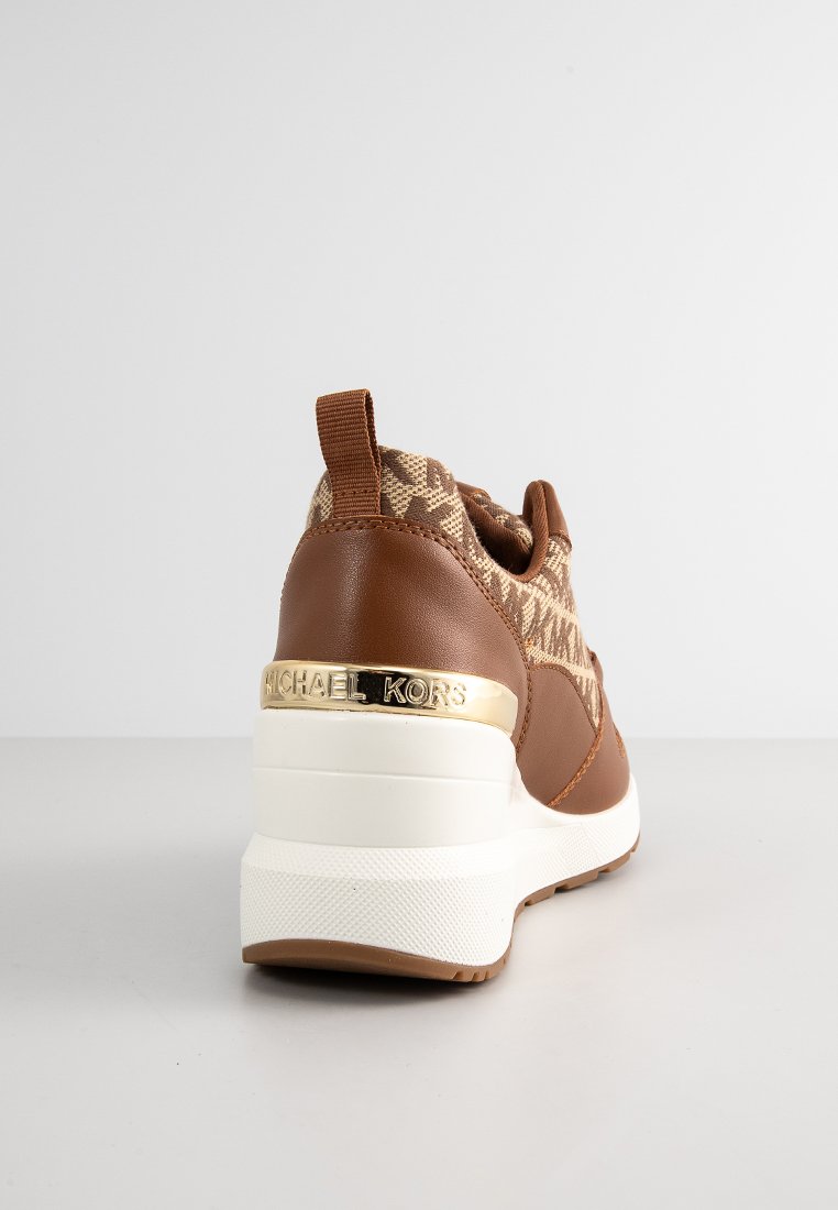 Zapatilla marrón con parte superior de tela que presenta un diseño estampado, detalles en cuero, una suela blanca elevada y un logo dorado en el talón.