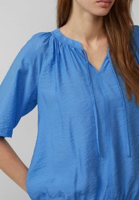 Blusa leggera blu con scollo a V, maniche corte, colletto arricciato e dettagli con coulisse sul davanti. Texture morbida e stropicciata.