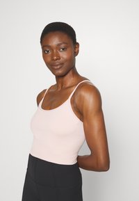Top sem mangas canelado e ajustado em tecido rosa claro, com alças finas e decote redondo. Combinando com leggings pretas de cintura alta.