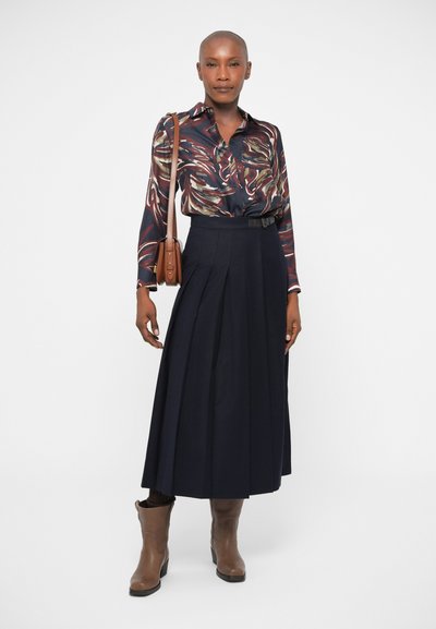Langærmet, flerfarvet mønstret bluse kombineret med en marineblå, plisseret nederdel. Brune ankelstøvler og en lille brun skuldertaske fuldender looket.