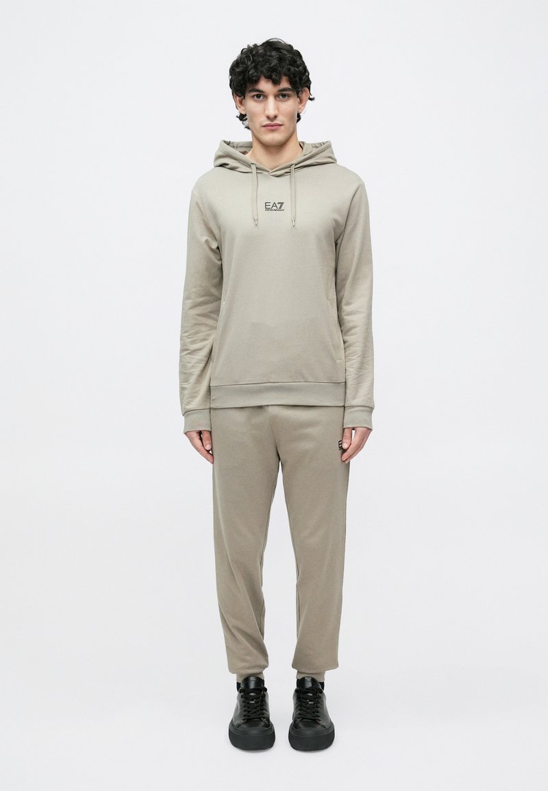 Béžová mikina s kapucňou a zladené zúžené tepláky z mäkkej látky. Mikina má šnúrky na kapucni, logo na hrudi a rebrované manžety. Čierne topánky.