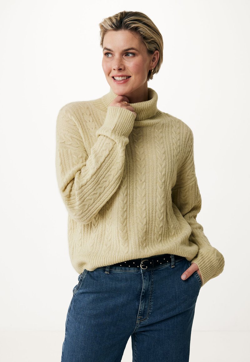 Maglione lavorato a maglia in morbido beige con colletto alto, caratterizzato da motivi a cavo testurizzati. Abbinato a jeans in denim blu e una cintura nera con borchie.