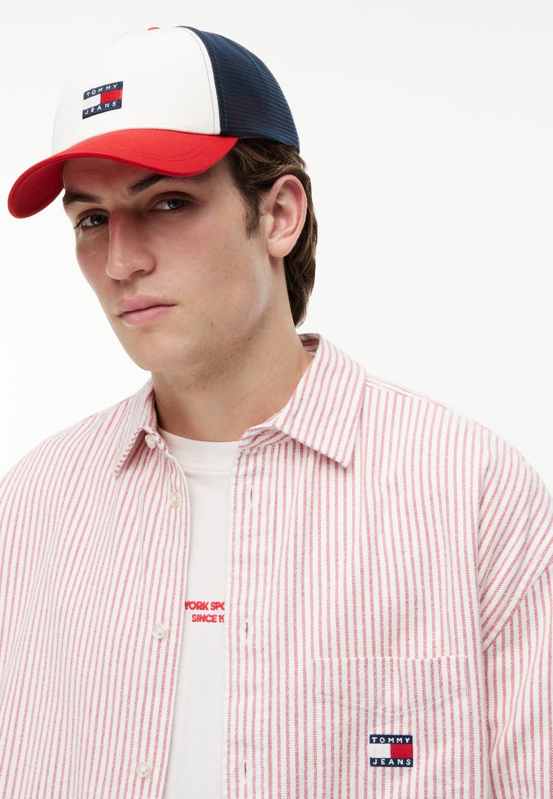Jeune homme portant une casquette Tommy Jeans blanche, rouge et marine, un t-shirt blanc avec un texte rouge, et une chemise à rayures rouges et blanches avec un écusson Tommy Jeans.