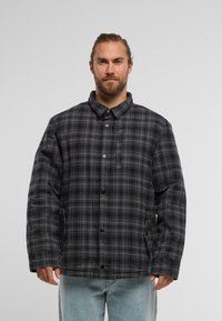 Melnā un pelēkā laukuma raksta jaka ar pogu aizdari, stāvošu apkakli un sānu kabatām. Iezīmē brīvu piegriezumu un quilted tekstūru.