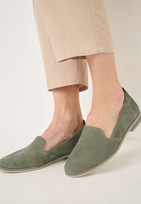 Groene suède instapschoenen met een lage hak, voorzien van een V-vormige opening en een gladde textuur, gecombineerd met lichtbeige cropped broeken.
