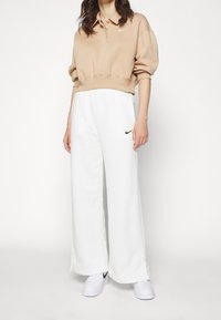 Beige, kurzes Sweatshirt mit Kragen und Nike-Logo, kombiniert mit weißen, weiten Sweatpants, die ein schwarzes Nike-Logo aufweisen. Weiße Sneakers.