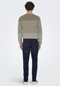 Olivgrön tröja med ribbad textur, med en gradientdesign. Bärs med mörkblå jeans och vita sneakers. Avslappnad passform.