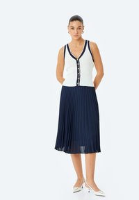 Witte geribbelde tanktop met marineblauwe bies en knoopjes, gecombineerd met een marineblauwe geplooide midi-rok. Model draagt witte pumps met spitse neuzen.