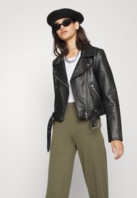 Casaquinho de motoqueiro em couro preto com fechos de correr prateados, design assimétrico e detalhe de cinto ajustável, usado sobre uma blusa branca e calças verdes.