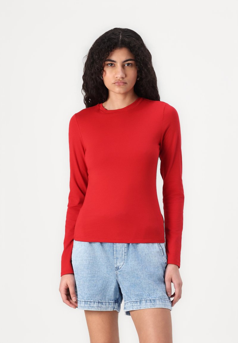 Cămașă roșie cu mâneci lungi, realizată dintr-un material neted, cu decolteu rotund. Asortată cu pantaloni scurți din denim bleu deschis, cu o croială lejeră.