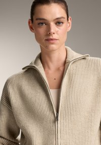 Beige gerippter Zip-Up-Pullover aus strukturiertem Stoff. Verfügt über einen Stehkragen und einen metallischen Reißverschluss. Trägt man über einem taillierten, neutralen Oberteil.