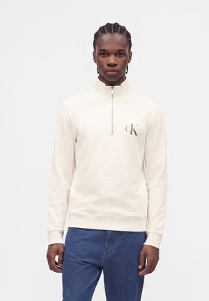 Joven con trenzas, vestido con una sudadera crema con cremallera hasta el pecho y logo de CK, y vaqueros azules, de pie frente a un fondo blanco liso.