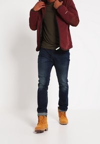 Kastanjefärgad vadderad jacka över en mörkgrön skjorta, i kombination med blå jeans och beige stövlar. Jackan har en dragkedja och svart fleecefoder.