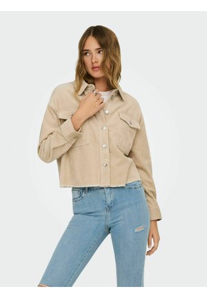 Jeune femme portant une veste courte en velours côtelé beige et un jean skinny bleu clair déchiré, posant avec la main près du col.