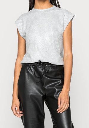 Mujer con una camiseta sin mangas de canalé gris claro y pantalones negros de piel sintética con cordón, sobre un fondo liso.