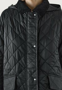 Zwarte doorgestikte jas met een ribfluwelen kraag, drukknopen en zijzakken. Het materiaal is glanzend en heeft een gestructureerd ruitpatroon.