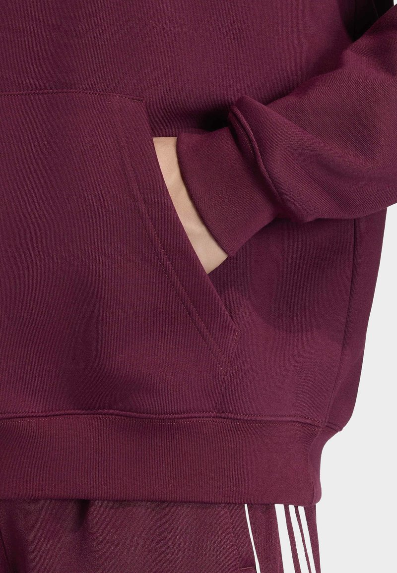 Mano appoggiata nella tasca anteriore di una felpa bordeaux con polsini a coste, abbinata a pantaloni bordeaux con strisce bianche.