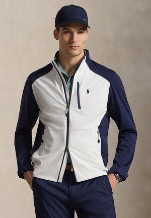 Férfi sötétkék sapkával, fehér és sötétkék cipzáras dzsekivel zöld csíkos galléros póló fölött, sötétkék nadrágban, kezével zsebében.